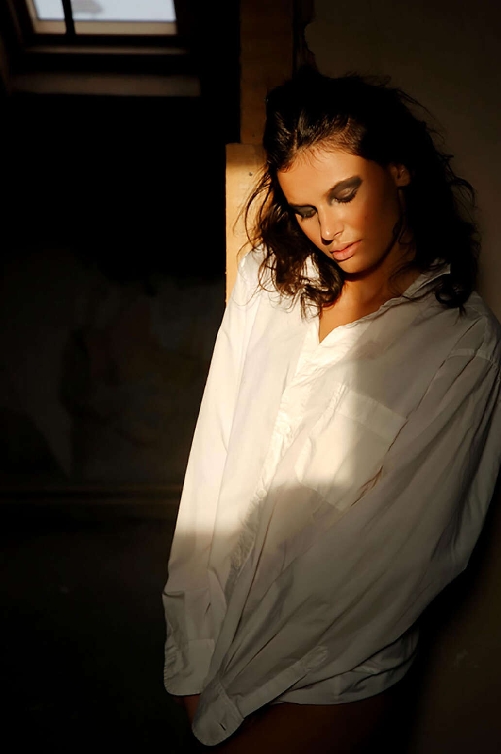 Portret artistic în lumină caldă, femeie în cămașă albă, sprijinită de perete, atmosferă melancolică și intimă