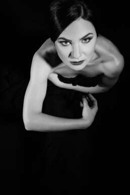 Portret artistic alb-negru al unei femei privite de sus, cu expresie intensă, în rochie neagră, iluminare dramatică, stil editorial