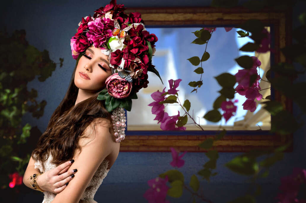Portret artistic al unei tinere femei purtând o coroană extravagantă din flori, fluturi și perle, în decor floral, cu fundal pictural