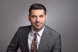 Portret profesional al unui bărbat tânăr, îmbrăcat în costum gri cu cravată paisley roșu-alb-albastru, privire directă și expresie calmă, fundal neutru.