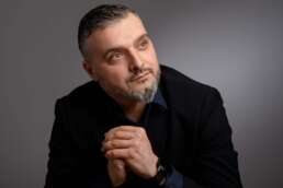 Portret profesionist al unui avocat cu barbă grizonantă, îmbrăcat în sacou închis la culoare, privind în lateral cu o expresie calmă și gânditoare. Fundal neutru.