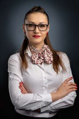 Portret profesional al unei femei cu ochelari, păr prins, cămașă albă și eșarfă imprimată, pe fundal întunecat. Poartă ruj roșu și o expresie încrezătoare.