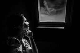 Portret alb-negru al miresei surprinsă în lumină naturală, privind pe fereastră, reflexie delicată în sticlă