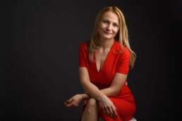 Femeie blondă, îmbrăcată într-o rochie roșie, stând relaxată pe un scaun, zâmbind cald, pe fundal închis.