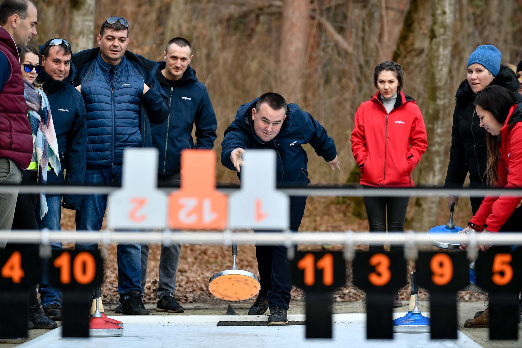 Angajați participă la o competiție de tip curling în aer liber, în cadrul unui eveniment de team building. Un coleg în prim-plan aruncă discul, în timp ce ceilalți urmăresc concentrați.