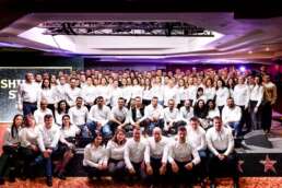 Poză de grup cu peste 100 de angajați îmbrăcați în cămăși albe, fotografiați la finalul unei conferințe corporate, pe o scenă decorată cu tematică “Shining Stars”