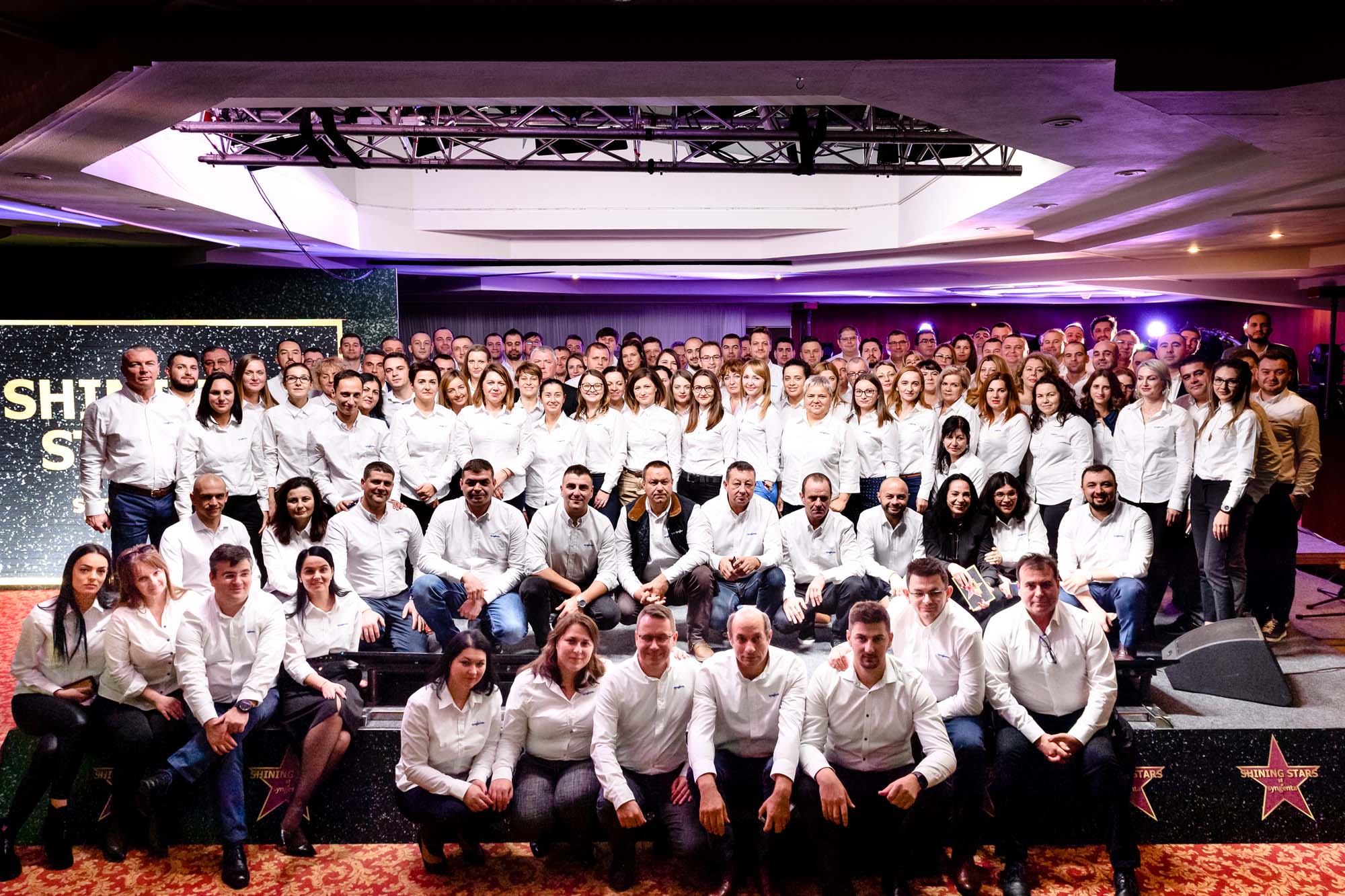 Poză de grup cu peste 100 de angajați îmbrăcați în cămăși albe, fotografiați la finalul unei conferințe corporate, pe o scenă decorată cu tematică “Shining Stars”