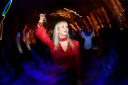 Femeie blondă dansează la o petrecere corporate, îmbrăcată într-o bluză roșie, înconjurată de lumini dinamice și participanți în fundal