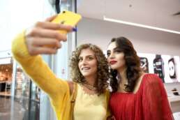 Două tinere fac un selfie cu un iPhone galben, la lansarea unui nou model iPhone în magazinul iSTYLE Băneasa, înconjurate de afișe cu smartwatch-uri