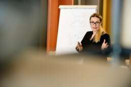 Trainer explicând în fața unui flipchart cu o diagramă, vorbind cu expresivitate și implicare