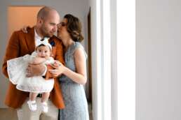 Fotograf botez București – fetiță în rochiță albă ținută de tată, mamă zâmbitoare în fundal, scenă caldă de familie