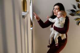 Mamă și copil în brațe, amândoi arătând cu degetul spre fereastră, îmbrăcați elegant – moment de conexiune și descoperire în ziua botezului